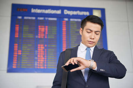 Portrait of handsome Asian businessman checking time on wristwatch while waiting for his flight in airport, standing against departure boardの写真素材