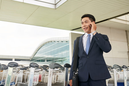 Portrait of modern Asian businessman speaking by phone leaving airport with luggage and smilingの写真素材