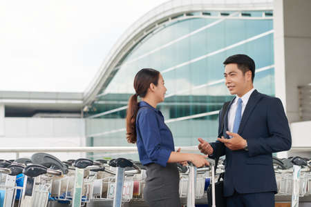 Portrait of modern Asian business partners, man and woman, talking in airport, smiling and gesturing activelyの写真素材