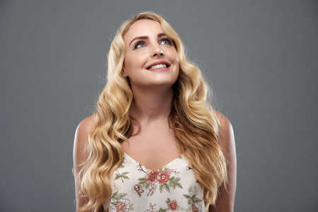Portrait of pretty young woman with wavy blond hair looking up and smiling against grey backgroundの写真素材