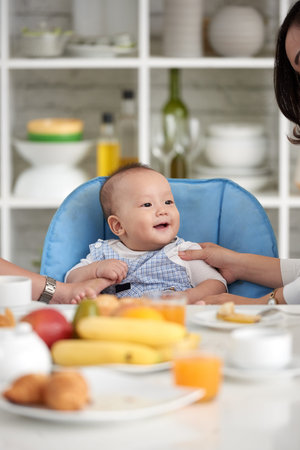 Portrait of adorable Asian baby boy smiling, sitting in little chair with family at dining table...