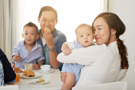 Happy Asian woman cuddling with cute baby boy at breakfast table with family on sunny morningの写真素材