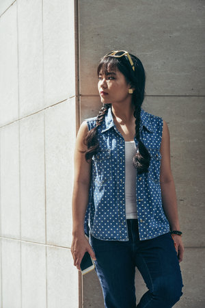 Beautiful Asian woman looking into distance thoughtfully while leaning on outer wall of modern...