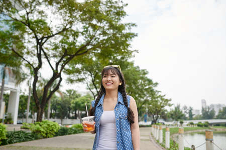 Smiling Vietnamese woman with plastic cup of iced tea in hand enjoying summer day and picturesque view on riverwalk, waist-up portraitの写真素材