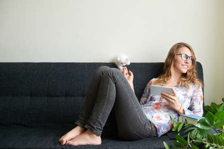 Smiling young woman in glasses lying on sofa with smartphone and playing with her catの写真素材