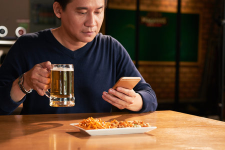 Man drinking beer and looking at smartphoneの写真素材