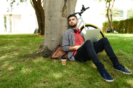 Pensive young man sitting on the grass under the tree when laptop on his knees