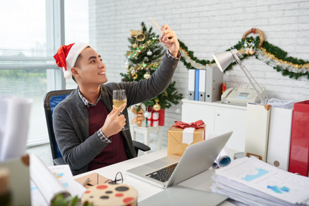 Asian businessman in Santa hat taking Christmas selfie with champagne glass at workplaceの写真素材