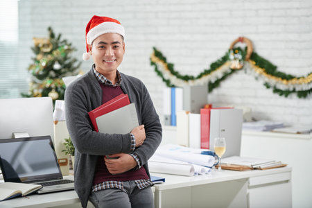 Portrait of handsome Asian man in Santa hat posing at workplace in office decorated for Christmasの写真素材