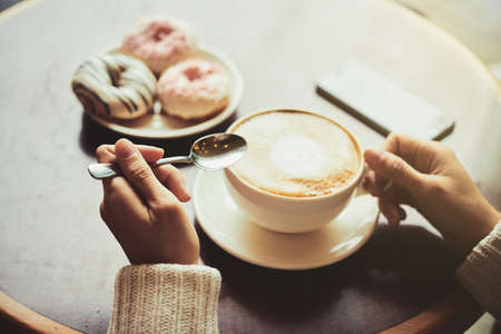 Unrecognizable young woman wearing knitted cardigan enjoying fragrant cappuccino and delicious doughnuts while sitting at cozy small cafeの写真素材