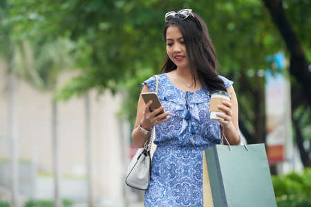 Fashionable Asian woman walking down the street with shopping bags and using smartphoneの写真素材
