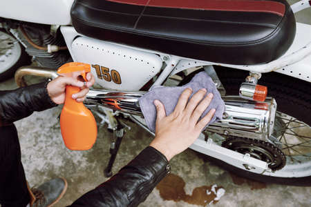 Close-up shot of unrecognizable man sitting on haunches and wiping dirt from his motorcycle with help of rag and cleaning sprayの写真素材