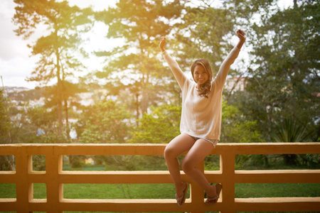 Happy Asian woman sitting on balcony railing in resort hotel and smiling at cameraの写真素材