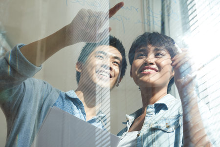 Smiling young Asian developers writing project plan on glass wall