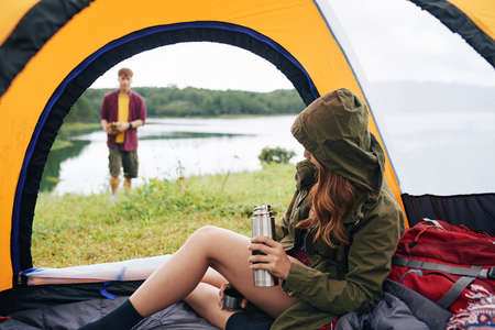 Young woman sitting in tent and drinking tea when her boyfriend is making campfireの写真素材
