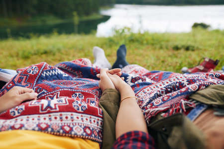 Couple of travelers holding hands when lying under blanket by the lakeの写真素材
