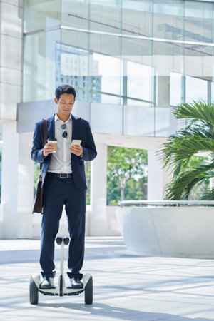 Young Vietnamese businessman with take-out coffee and smartphone standing on gyroscooterの写真素材