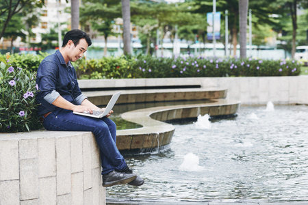 Vietnamese young man sitting on the edge of fountain in park and working on laptopの写真素材