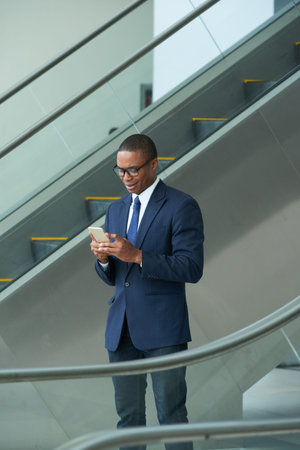 African-American businessman standing on escalator and reading messaged in his smartphoneの写真素材