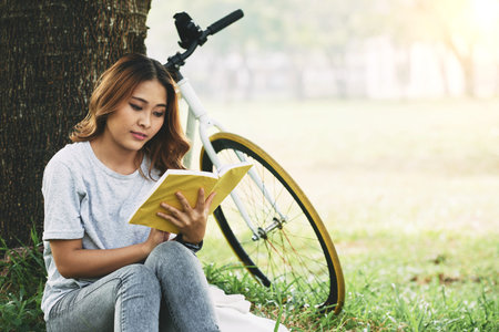 Pretty Vietnmamese young woman sitting under the tree and reading an interesting book