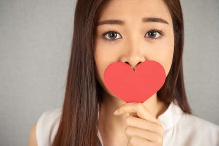 Head and shoulders portrait of attractive Asian woman looking at camera with deep gray eyes while covering her mouth with heart shaped piece of paperの写真素材