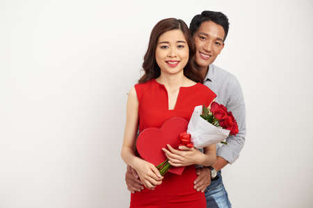 Group portrait of cheerful Asian couple looking at camera with wide smiles while celebrating Valentines Day together, pretty woman holding bouquet of red roses and heart shaped gift box in handsの写真素材