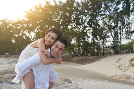 Enjoying vacation at exotic resort: handsome Asian man with wide smile looking away while giving piggyback ride to his pretty girlfriendの写真素材