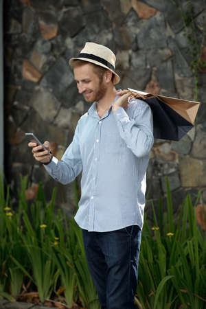 Handsome young man texting on smartphone and holding shopping bags outdoorsの写真素材