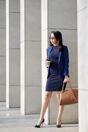 Full length portrait of attractive young entrepreneur wearing sunglasses standing at facade of office building, drinking coffee from paper cup and enjoying fresh airの写真素材