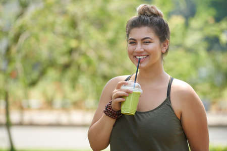 Smiling young woman wearing top looking at camera while drinking smoothie from straw and enjoying picturesque view at green public parkの写真素材