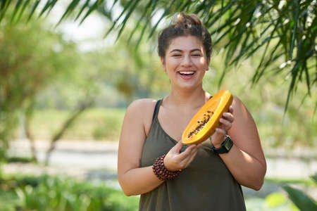 Joyful young woman posing for photography with toothy smile while holding ripe papaya in hands, sunny green park on backgroundの写真素材