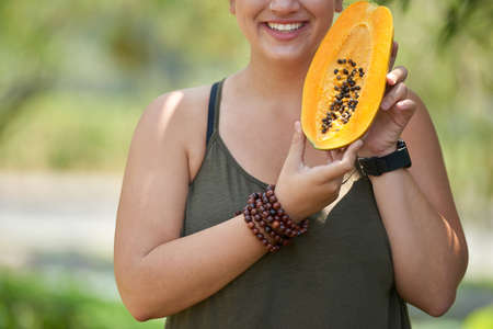 Unrecognizable young woman with charming smile standing at green public park illuminated with sunbeams and holding ripe papaya in handsの写真素材