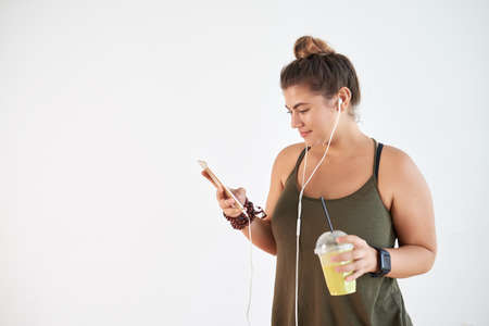 Pretty young woman listening to music with help of smartphone and refreshing herself with smoothie while standing against white background, copy spaceの写真素材