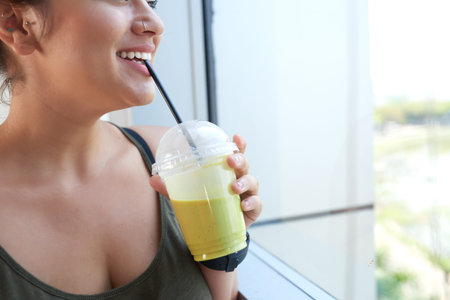 Close-up shot of cheerful young woman looking through window while refreshing herself with delicious smoothieの写真素材