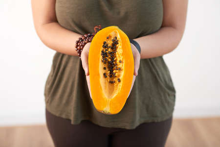 Close-up shot of obese young woman wearing top and leggings standing against white wall and holding ripe papaya in handsの写真素材