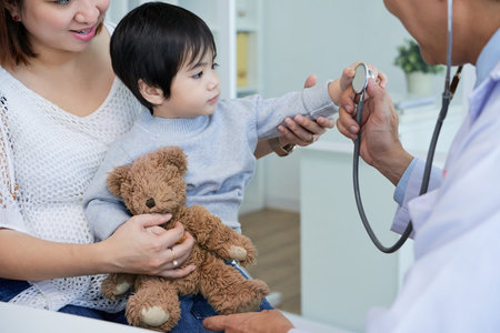 Curious Asian boy sitting on laps of his pretty mother and examining stethoscope with interest while having consultation at pediatrician officeの写真素材