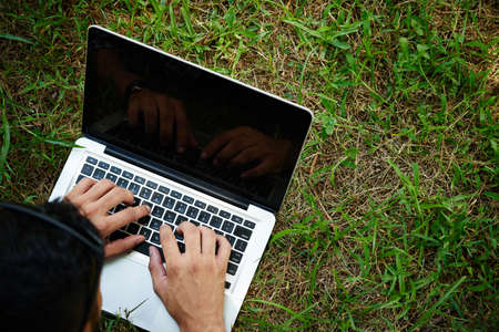 Close-up shot of unrecognizable man lying on green lawn and using modern laptop while spending weekend outdoorsの写真素材