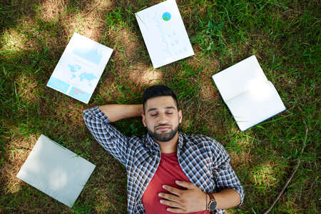 Handsome bearded manager lying on green lawn with closed eyes while taking break from work, documents and notepad illuminated with sunbeamsの写真素材