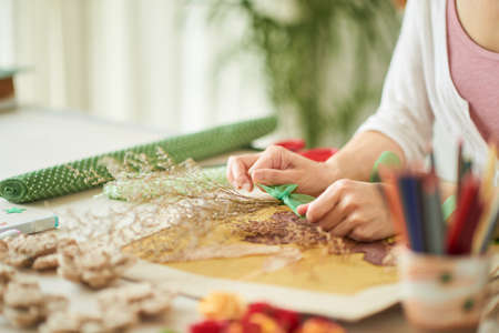 Close-up shot of talented young designer in casualwear making creative decor item for her client while sitting at desk, interior of modern studio on backgroundの写真素材