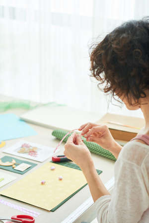 Profile view of curly woman in casualwear sitting at wooden table while enjoying process of handmade greeting card creationの写真素材