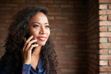 Head and shoulders portrait of attractive Asian woman looking away with wide smile while talking to friend on smartphone, brick wall on backgroundの写真素材