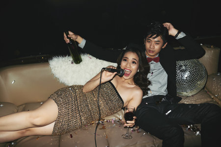Hanging out at nightclub: handsome Asian man with wine bottle sitting on cozy sofa and looking at camera while his attractive young girlfriend singing karaokeの写真素材