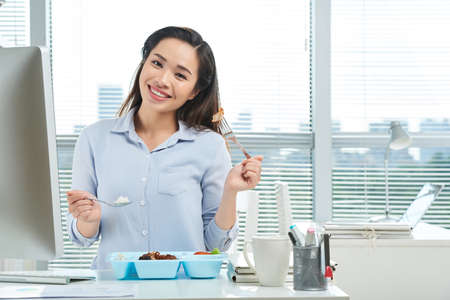 Pretty smiling business lady eating lunch at her office tableの写真素材