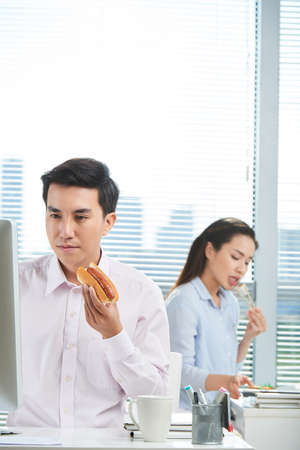 Young Vietnamese entrepreneur eating hot dog and working on computer in officeの写真素材