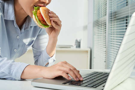 Female entreprepreneur eating chicken burger when working on laptop in officeの写真素材