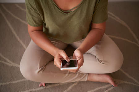 Unrecognizable woman wearing trousers and T-shirt sitting in lotus position and browsing Internet with help of modern smartphone, close-up shotの写真素材