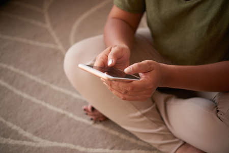 Unrecognizable woman sitting in louts position on carpet and browsing Internet on modern smartphone, close-up shotの写真素材