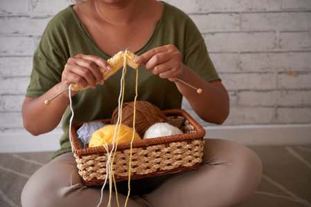 Woman sitting in lotus position against white brick wall and wrapped up in knitting colorful...