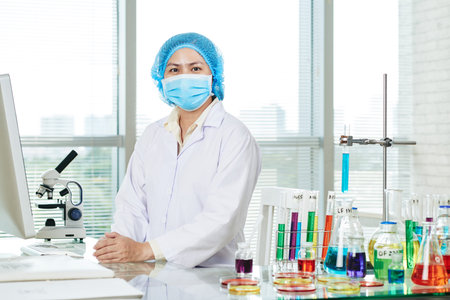 Portrait shot of confident Asian microbiologist wearing white coat and medical mask sitting at desk...