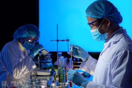 Gifted Asian scientist wearing rubber gloves and safety masks working together on creation of cancer vaccine, interior of dark laboratory on backgroundの写真素材